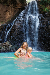 In tropical jungle happy mother with kids stand under waterfall, explore rainforest nature. Travel adventure, hiking activity with child. Lifestyle on family summer vacation and weekend walking tour