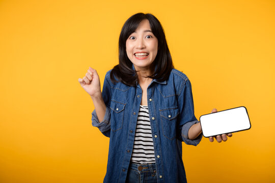 Portrait beautiful young asian woman happy smile dressed in denim jacket showing smartphone screen isolate on yellow studio background. New smartphone application concept