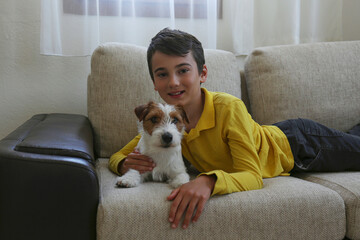 Portrait of 11 year old boy playing with wire haired jack russell terrier puppy at home. Tween boy with rough coated pup on the couch. Close up, copy space, background.
