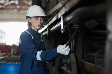 Female engineer inspecting quality parts of locomotive engine, wearing safety uniform, helmet and gloves in locomotive garage. Female railway technician worker repair train wheel in garage workshop