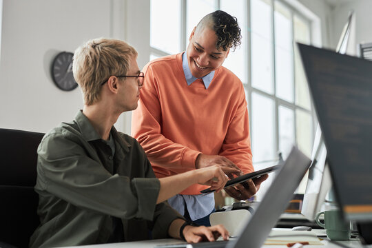 Smiling young colleague consulting with programmer about new software on digital tablet during their work in office