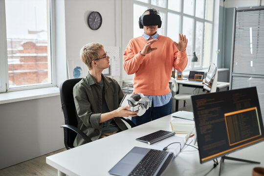 Young programmer testing new software in VR glasses together with his colleague during teamwork in IT office