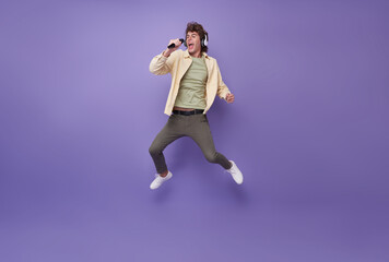 Full body crazy hipster guy jumping high holding microphone singing music favorite song isolated on purple studio background.