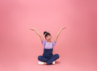 happy little asian girl sitting and presenting or showing open hand palm with copy space for product isolated on pink copy space background.