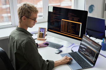 Rear view of young programmer in eyeglasses writing codes on different gadgets while working at his...