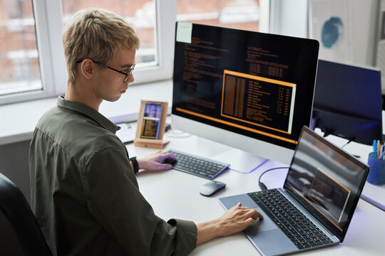 Rear view of young programmer in eyeglasses writing codes on different gadgets while working at his workplace in IT office