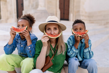 Multiracial kids with mother eating watermelon in street during hot sunny days, summer holiday travelling concept.