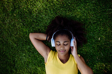 Multiracial girl lying down in grass and enjoying music in headphones, top view.