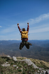 Concept of travel and healthy, active lifestyle. Young guy with dreadlocks went hiking in mountains. Man with yellow backpack jumps up on top of hill and raised two hands up. Rear view.