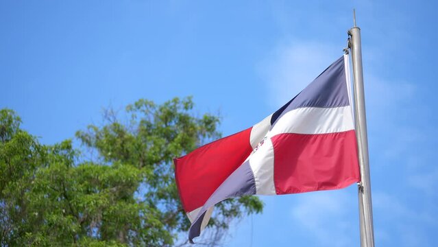 National flag of the Dominican Republic on flagpole waving in the wind with blue sky and green tree on the background Generative AI