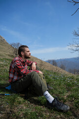 Portrait of young guy with dreadlocks and beard against mountains and blue sky. Young male tourist on hike resting sitting on clearing among primroses. Concept of travel and active lifestyle.