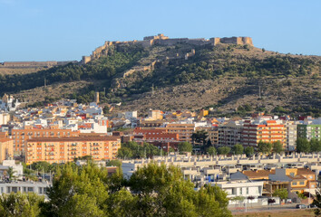 Fototapeta na wymiar Buildings and houses in city, aerial view. Sagunto Castle on mountain. Ruins walls of Fortress Castle at town of Sagunto. Fortress Castillo in Mountains hills. Houses roofs in town at mountains.