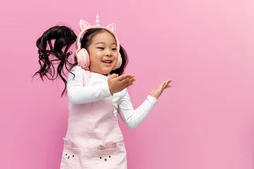 Fotobehang Dansschool asian girl in pink children's headphones and glasses listens to music and dances on pink isolated background