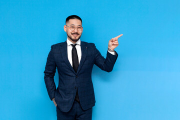 successful asian businessman in suit pointing with hand at copy space on blue isolated background