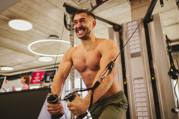 A strong sportsman is exercising on a cable machine with effort in a gym.