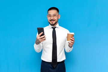 successful asian businessman in white shirt uses smartphone and holds paper cup of coffee on blue isolated background