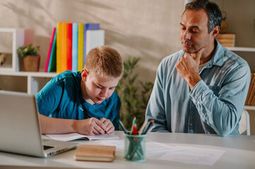 Blond teenage boy writing his homework while his father is sitting next to him.