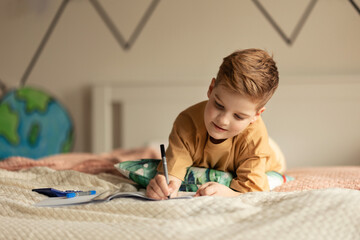 Little boy lying on a bed and doing his homework.