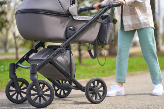 Cropped view of young mom pushing baby stroller while strolling the city park street in spring day