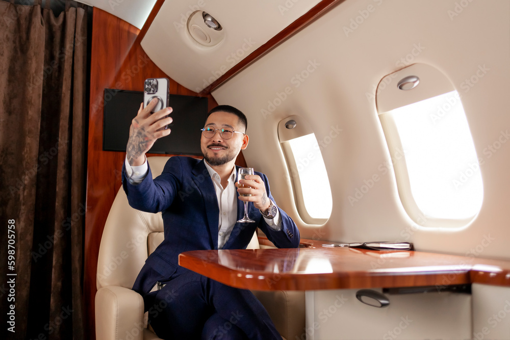 Wall mural asian businessman in suit and glasses sits in private jet with glass of champagne and communicates via video call