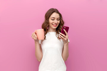 young cute woman in white t-shirt holds cup with drink and uses smartphone on pink isolated background