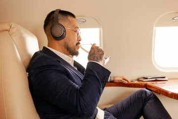 asian businessman in suit sits in private jet drinks champagne and listens to music on headphones