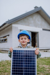 Portrait of little boy holding solar panel, in front of their new unfinished house.