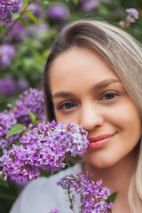 Fototapeta premium Portrait of beautiful young blue-eyed blonde woman near blooming lilac. Spring.