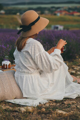 A woman in a white dress sits in the middle of a lavender field and holds fragrant tea in her hands. Blooming lavender. Picnic in Provence style.