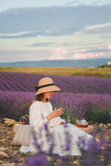 A woman in a white dress sits in the middle of a lavender field and holds fragrant tea in her hands. Blooming lavender. Picnic in Provence style.