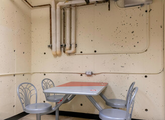 A grey and red steel table and four steel swivel chairs bolted to the floor in a room with rough yellow concrete walls, utility pipes, nobody