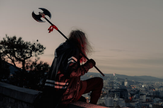 Young man long blond hair viking style with an ax in his hands, sitting above city on mountain top during sunset