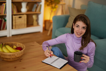 Pretty young woman sitting in cozy living room and making notes to her diary