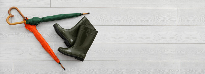 Umbrellas and rubber boots on light wooden background with space for text