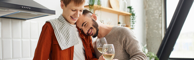 overjoyed bearded man leaning on redhead boyfriend while clinking wine glasses in kitchen, banner. 