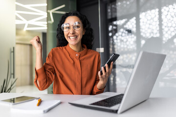 Portrait of successful Latin American business woman, worker in glasses and curly hair looking at camera and smiling happy celebrating triumph and winning success sitting at table holding phone.