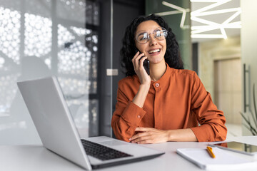Young beautiful businesswoman talking on the phone and looking towards the window, happy hispanic...