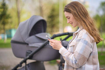 Side portrait of a Caucasian Red haired young pretty woman, happy mother using mobile phone while resting in a green park during stroll with her newborn child sleeping in the baby carriage