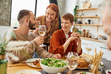 smiling woman toasting with wine near son with gay boyfriend near delicious meal in kitchen. 