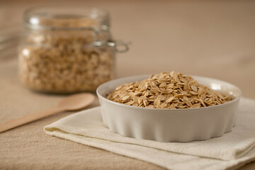 Oat flakes, cereal in a white ceramic bowl on a linen tablecloth on the table, a spoon and a glass jar with oatmeal