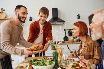 cheerful gay couple serving grilled chicken for family supper with parents in kitchen. 