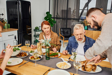 bearded gay man cutting grilled chicken during family dinner with boyfriend and parents in kitchen. 