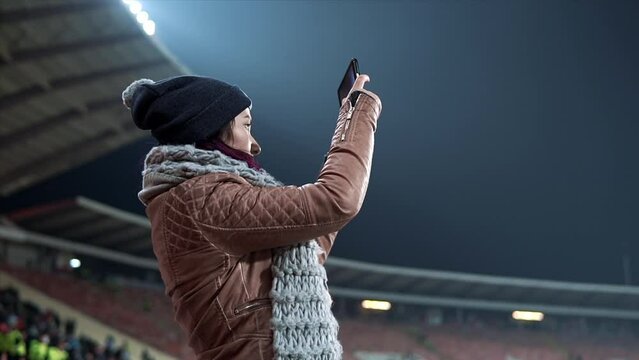 Woman taking photos or recording videos of live music concert with smartphones at open air festival or at sport game. Bright colorful stage lighting. Girl dressed in hat and scarf shooting video at