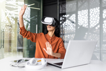 Young successful woman inside the office using vr glasses, virtual reality simulator cross charts and documents online, latin american woman using laptop at work.