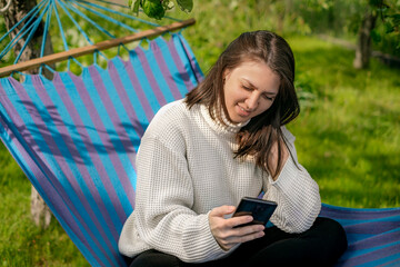 A woman is sitting on a hammock in the garden holding a phone in her hand and writing a message. Online communication using a smartphone.