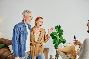 amazed woman showing wow gesture near gay couple with flowers and wine bottle at home. 