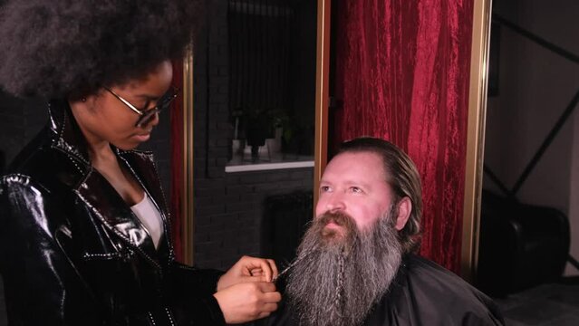 A stylish African-American woman in a black raincoat in a barber shop braids her braids into a beard for a male client.