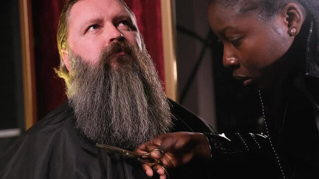 an African-American hairdresser girl in a black raincoat cuts a man's beard with scissors.