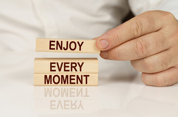 In the hands of a person and on a reflective surface are wooden blocks with the inscription - Enjoy Every Moment