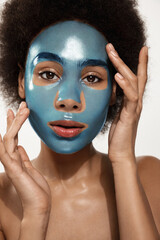 Headshot of good looking dark skinned woman with curly hairstyle, focused aside, has white clay mask on face, prepares for date with guy, does beauty treatments, isolated over blue background